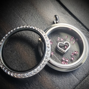 May include: A silver locket necklace with a clear glass insert. The locket is decorated with a row of small clear crystals around the edge. Inside the locket, there are pink crystals scattered around a silver heart charm.