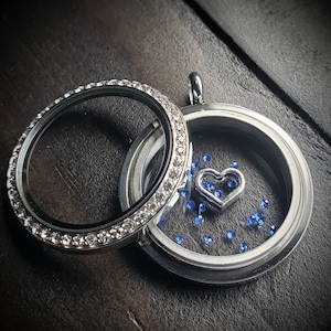 May include: A silver locket with a clear glass insert and a small silver heart charm inside. The locket is surrounded by a row of clear rhinestones and is filled with tiny blue rhinestones.