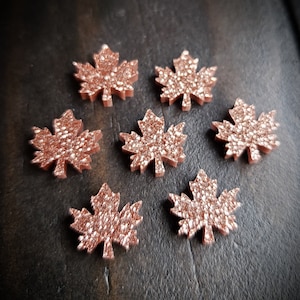May include: Seven rose gold glitter maple leaf embellishments. The leaves are made of a sparkly material and have a slightly raised texture.