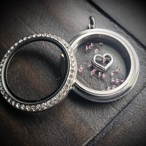 May include: A silver locket with a clear glass insert. The locket is decorated with a row of clear rhinestones around the edge. Inside the locket, there are small pink crystals and a silver heart charm.