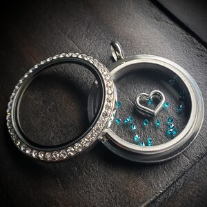 May include: A silver locket with a clear glass insert and a small silver heart charm. The locket is surrounded by a row of clear rhinestones and is filled with turquoise glitter.