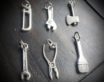 May include: A set of seven silver charm pendants shaped like various tools, including a wrench, pliers, a saw, an axe, a paintbrush, and two wrenches.