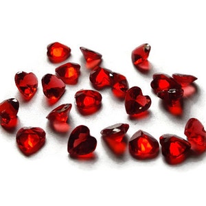 May include: A scattering of small, red, heart-shaped glass gems on a white background.