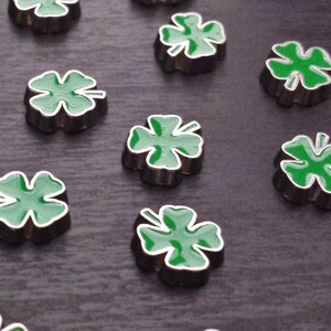 May include: A collection of 14 small, silver-colored four-leaf clover charms with green enamel. The charms are arranged in a pattern on a dark surface.