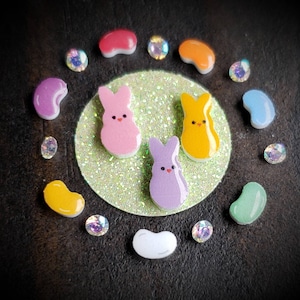 May include: A close-up of a colorful Easter craft project.  The craft features three pastel-colored bunny shapes, yellow, pink, and purple, surrounded by colorful jelly beans and rhinestones.