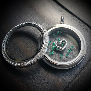 May include: A silver locket necklace with a clear glass insert. The locket has a silver heart charm inside with green glitter surrounding it.