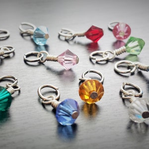 May include: Assortment of colorful glass beads attached to silver rings. The beads are in various shapes and sizes, including round, square, and diamond-shaped. The beads are in shades of blue, green, red, pink, and clear.