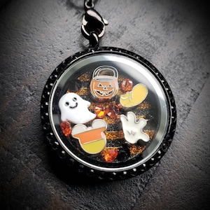 May include: Black locket charm with a Halloween theme. The charm features a ghost, a pumpkin, a candy corn, a Mickey Mouse ear, and a hand. The charm is filled with orange glitter and rhinestones.