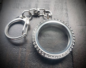 May include: A silver locket keychain with a clear glass insert and a silver chain. The locket is decorated with a row of small, clear rhinestones around the edge.