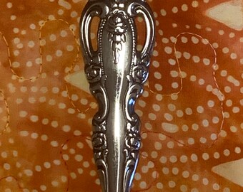 May include: Close-up of the ornate handle of a silver spoon. The handle is decorated with a floral design and has two cut-out ovals.
