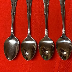 May include: Four silver spoons with ornate handles and a polished finish. The spoons are arranged in a row on a red surface.