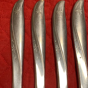 May include: Four stainless steel knives with a starburst design on the handle. The knives have a curved blade and a decorative etched pattern. The background is a red quilted fabric.