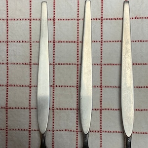 Puede incluir: Tres tenedores de plata con mangos redondeados y dientes puntiagudos, dispuestos verticalmente sobre un mantel a cuadros blanco y rojo.