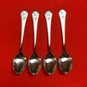 May include: Four silver teaspoons with a floral design on the handle. The spoons are arranged in a row on a red surface.