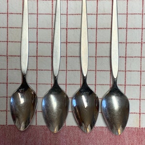 May include: Four silver spoons with a simple, elegant design are arranged on a red and white checkered tablecloth. Each spoon has a long, slender handle and a rounded bowl. The spoons are made of a shiny, reflective metal.