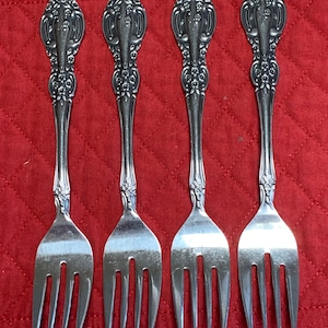 May include: Four silver forks with ornate floral and scrollwork detailing on the handles. The forks are arranged in a row against a red quilted background, showcasing their elegant design.