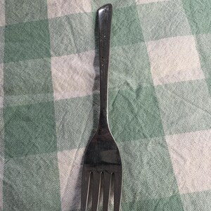 May include: A stainless steel fork with four tines, resting on a green and white checkered tablecloth. The handle has a decorative pattern. The fork is a common utensil for eating.