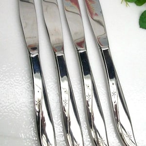 May include: Set of four stainless steel dinner knives. Each knife has a curved handle with a starburst design and a polished blade. The word "STAINLESS" is visible on one blade.