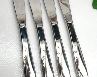 May include: Set of four stainless steel dinner knives. Each knife has a curved handle with a starburst design and a polished blade. The word "STAINLESS" is visible on one blade.