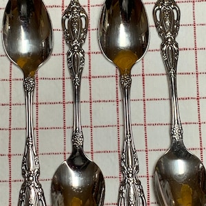 May include: Four silver spoons with ornate, detailed handles and oval bowls. The spoons are arranged on a red and white checkered surface, showcasing their reflective, polished surfaces. The handles feature floral and scrollwork designs.