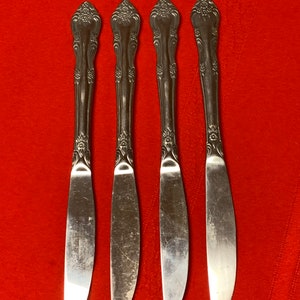 May include: Four silver dinner knives with ornate handles and a floral design. The knives are arranged in a row on a red surface.