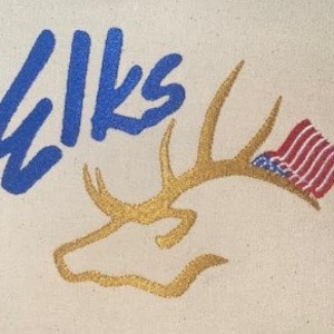 Puede incluir: Una bolsa de lona beige bordada con una cabeza de alce dorada con cuernos y una bandera estadounidense. La palabra "WIKS" está bordada en azul en la bolsa.