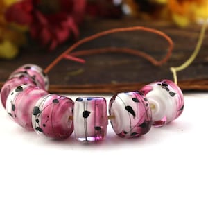 May include: A strand of pink and white glass beads with black abstract designs. The beads are cylindrical and have a glossy finish.