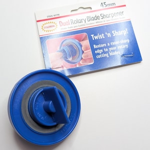 Könnte beinhalten: Ein blauer Kunststoff-Doppel-Rollklingenschärfer für 45-mm-Klingen. Die Verpackung enthält den Text Colonial Dual Rotary Blade Sharpener, Twist 'n Sharp! Restore a razor-sharp edge to your rotary cutting blades und die Nummer 5790.