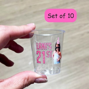 May include: Set of 10 clear plastic shot glasses with personalized pink and glitter stickers. The sticker features a photo of a person wearing a party hat and the text "LANI'S 21ST".