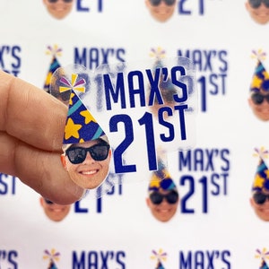 May include: A sheet of custom stickers featuring a repeating graphic of a person wearing a blue and yellow party hat and black sunglasses, accompanied by the blue text "MAX'S 21ST". One individual sticker is held up to show the clear adhesive design.