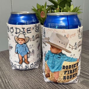 May include: Two white and blue can coolers with a photo of a baby boy wearing a cowboy hat and a blue shirt. The text on the coolers says "Bodie's First Rodeo".