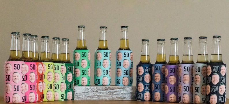 May include: A row of 15 colorful beer bottle coolers with a repeated pattern of a man's face and the number 50. The coolers are arranged in a row on a wooden surface.