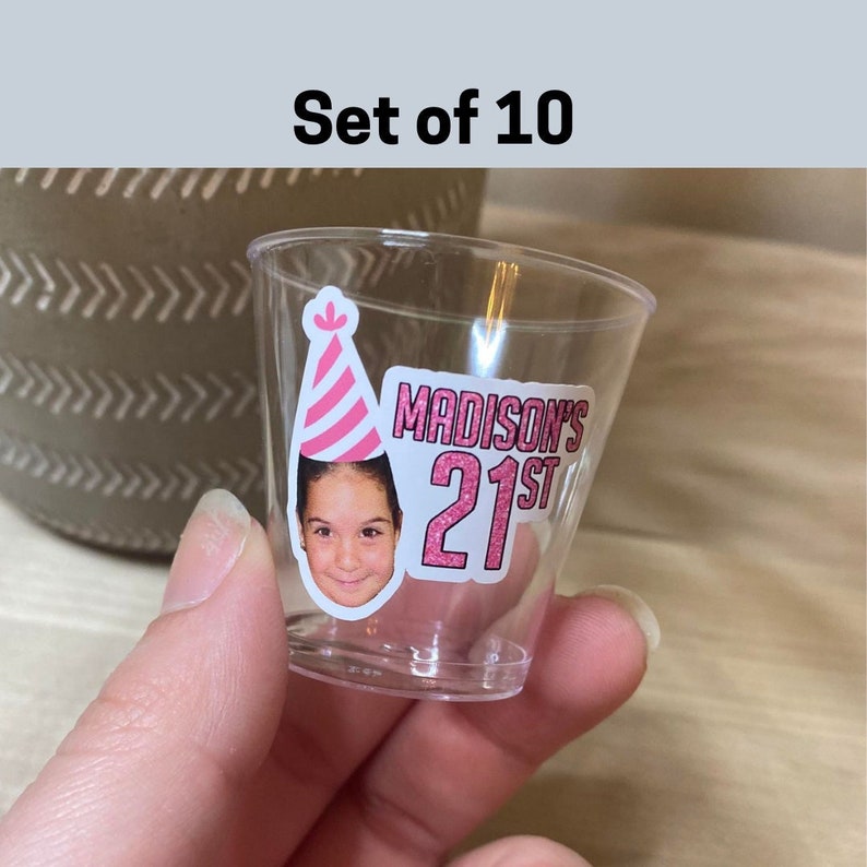 Może przedstawiać: Zestaw 10 przezroczystych plastikowych kieliszk&oacute;w do shot&oacute;w z personalizowanymi naklejkami. Każda naklejka zawiera r&oacute;żowy i biały kapelusz imprezowy, zdjęcie osoby i tekst "MADISON'S 21ST" w r&oacute;żowym brokacie.