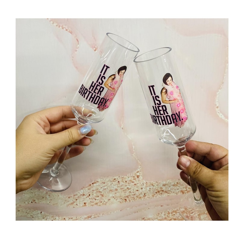May include: Two clear champagne flutes with a pink and black design that says "It is her birthday."  Each flute features a photo of a woman in a pink dress.