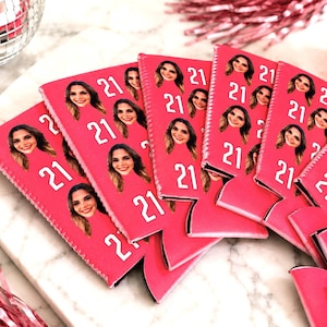 May include: A collection of pink can coolers, each featuring a photo of a woman and the number "21" in white. The coolers are arranged on a marble surface, with a disco ball and pink decorations in the background.