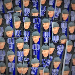 May include: A close-up of a stack of blue and black can coolers with the text "Old AF" printed on them. The coolers feature a repeated pattern of the text and a man's face.