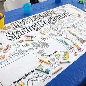 May include: A large white coloring sheet with the text "LPA D22026 Spring Regional" and illustrations of New York landmarks and food. A person is coloring the sheet with a yellow crayon. Crayons and a yellow cup are on the blue table.