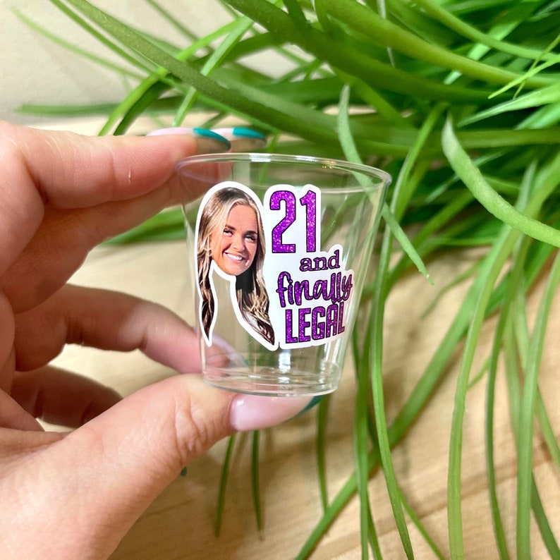 Może przedstawiać: Przezroczysty plastikowy kieliszek do shot&oacute;w z r&oacute;żowo-białą naklejką z napisem "21 and finally legal". Naklejka ma błyszczące wykończenie i zdjęcie twarzy kobiety.