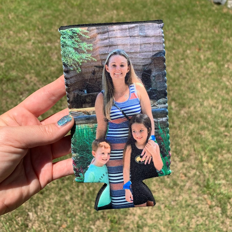 May include: A black and white can cooler with a photo of a family of three. The woman is wearing a blue and white striped dress and the two children are wearing a blue and white shirt and a black shirt with a Batman logo.