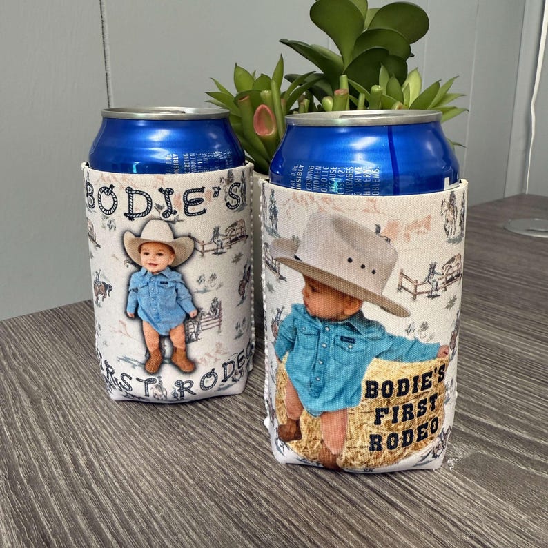 May include: Two white and blue can coolers with a photo of a baby boy wearing a cowboy hat and a blue shirt. The text on the coolers says "Bodie's First Rodeo".