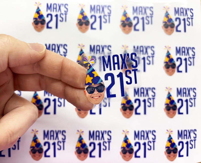 May include: A sheet of custom stickers featuring a repeating graphic of a person wearing a blue and yellow party hat and black sunglasses, accompanied by the blue text "MAX'S 21ST". One individual sticker is held up to show the clear adhesive design.