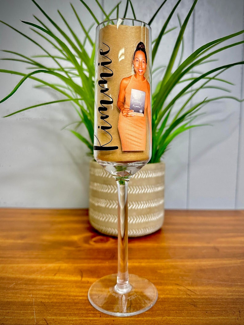 May include: A champagne flute with a gold-colored base and a clear stem. The flute has a personalized label with the name "Kimmie" and a photo of a woman holding a book. The flute is sitting on a wooden surface.