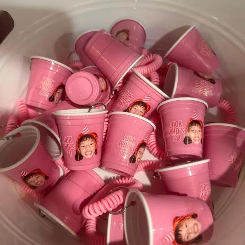 May include: A collection of pink plastic shot glasses, each featuring a printed photograph of a person wearing a party hat and the text "LOOK WHO'S 21!". Several of the cups are attached to pink coiled wristbands with metal key rings.