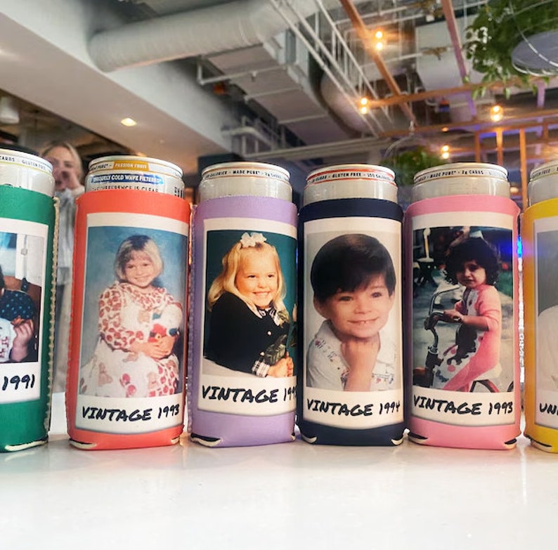 May include: A row of colorful foam beverage can coolers, each featuring a printed vintage photograph of a child and the text VINTAGE followed by a year. The coolers are green, orange, lavender, navy blue, pink, and yellow, holding metal cans.