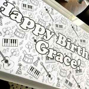 May include: A white banner with a black and white musical theme, featuring guitars, drums, piano keys, balloons, and musical notes. The words "Happy Birthday Grace!" are prominently displayed in large, bold letters.