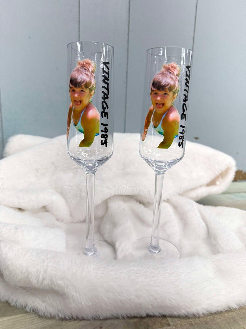 May include: Two clear glass champagne flutes with a vintage 1985 photo of a young girl in a swimsuit printed on each. The photo is of the same girl, but mirrored on each flute.