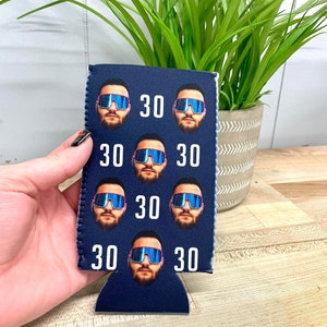 May include: A blue neoprene can cooler with a repeated pattern of a man's face wearing sunglasses and the number 30.
