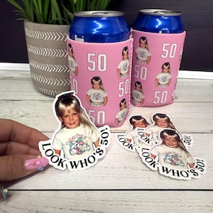 May include: Pink can coolers with a photo of a child and the number 50 printed on them. The text "LOOK WHO'S 50!" is on stickers with the same photo. A potted plant is in the background.