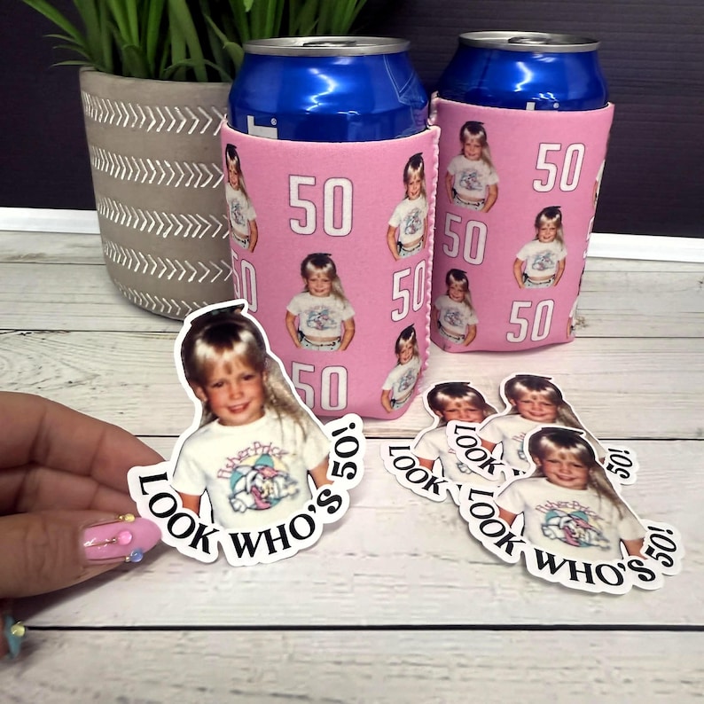 May include: Pink can coolers with a photo of a child and the number 50 printed on them. The text "LOOK WHO'S 50!" is on stickers with the same photo. A potted plant is in the background.