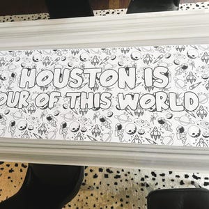 Może przedstawiać: Czarno-biała kolorowanka z motywem kosmicznym, przedstawiająca rakiety, planety i astronaut&oacute;w. Tekst "HOUSTON IS OUT OF THIS WORLD" jest wyraźnie widoczny. W rogu widoczne jest pudełko kredek.